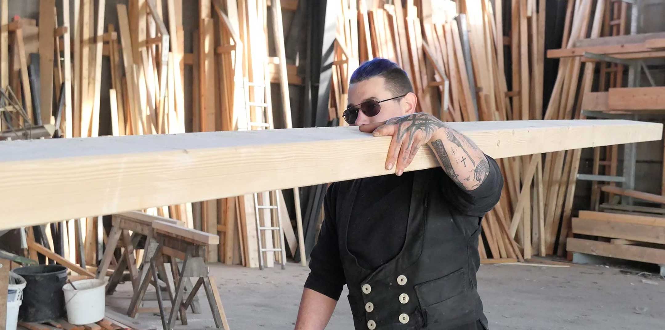 Eine Person in traditioneller Zunftkleidung trägt ein längeres Holzbrett auf der Schulter durch eine Werkstatt. Im Hintergrund sind aufrecht stehende Holzlatten und verschiedene Werkzeuge zu sehen.